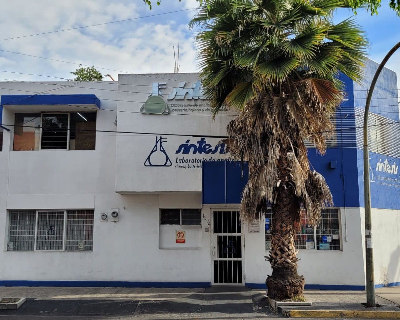 Fachada de Sintesis Laboratorio: edificio blanco de dos pisos con la torre azul del logotipo y una palmera al frente, en Ciprés 1904, Col. del Fresno, Guadalajara